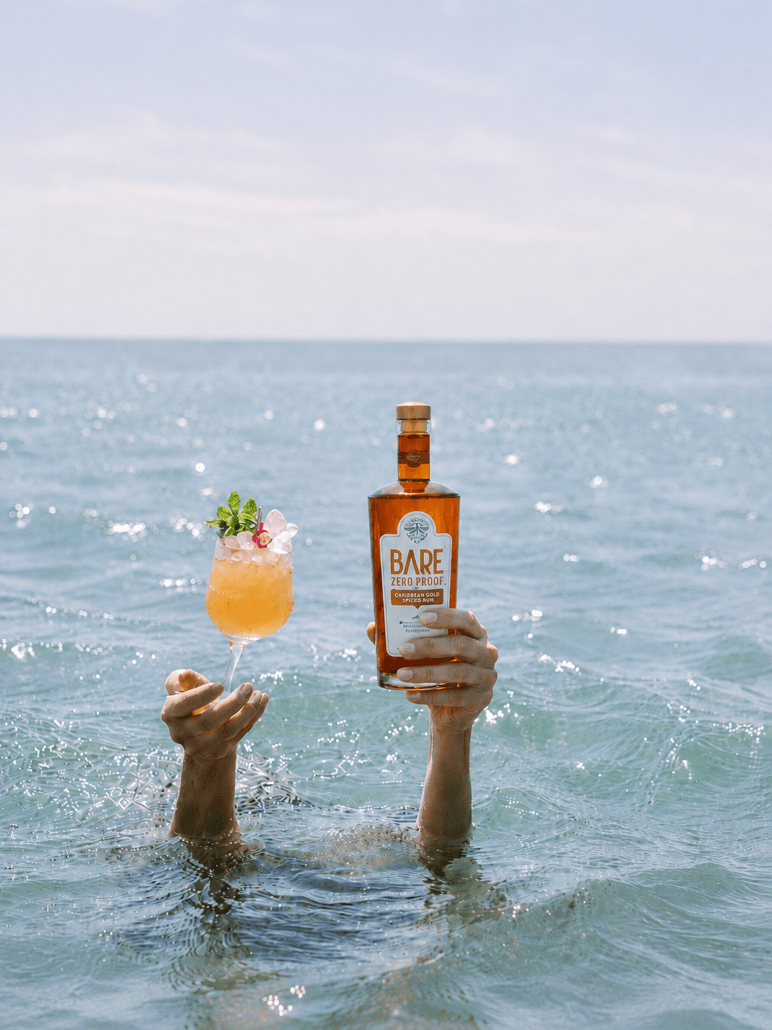 Two hands rising out of the sea, one holding a Mai Tai and the other a bottle of BARE Zero Proof spiced rum.